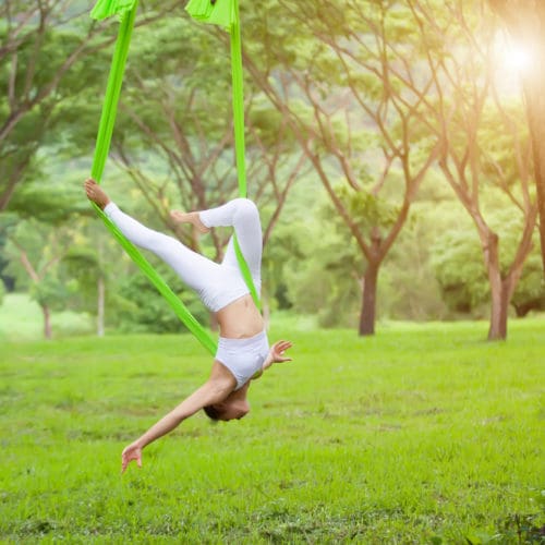 Person practising aerial yoga upside down in a green hammock outdoors, surrounded by trees and bright natural sunlight.