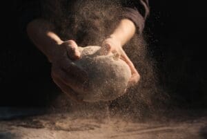 News & Blogs - Close-up of baker’s hands kneading dough with flour dust in the air.