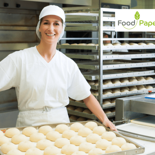 Sustainability in Baking - Baker in a kitchen holding a tray of dough balls with racks of fresh bread in the background.