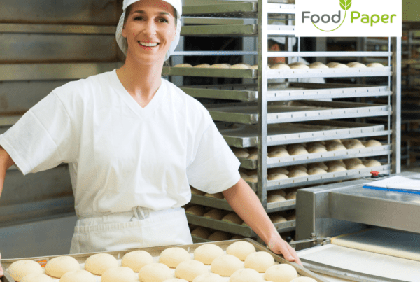 Baker in a kitchen holding a tray of dough balls with racks of fresh bread in the background.