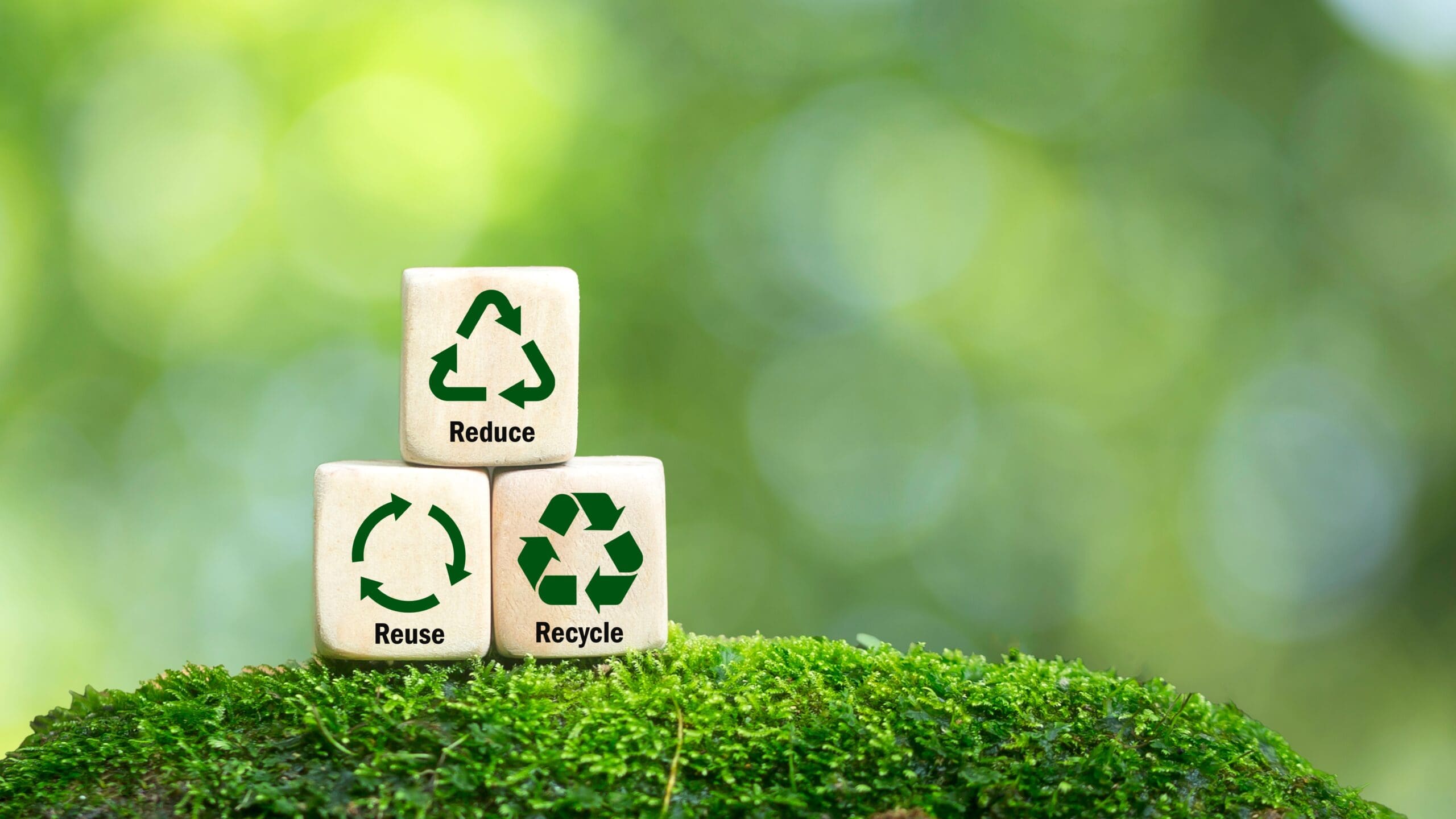 Sustainable Baker - Wooden blocks with reduce, reuse, and recycle symbols on moss with a blurred green background.