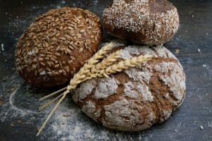 Three rustic bread loaves topped with seeds and flour beside wheat stalks on a wooden surface.